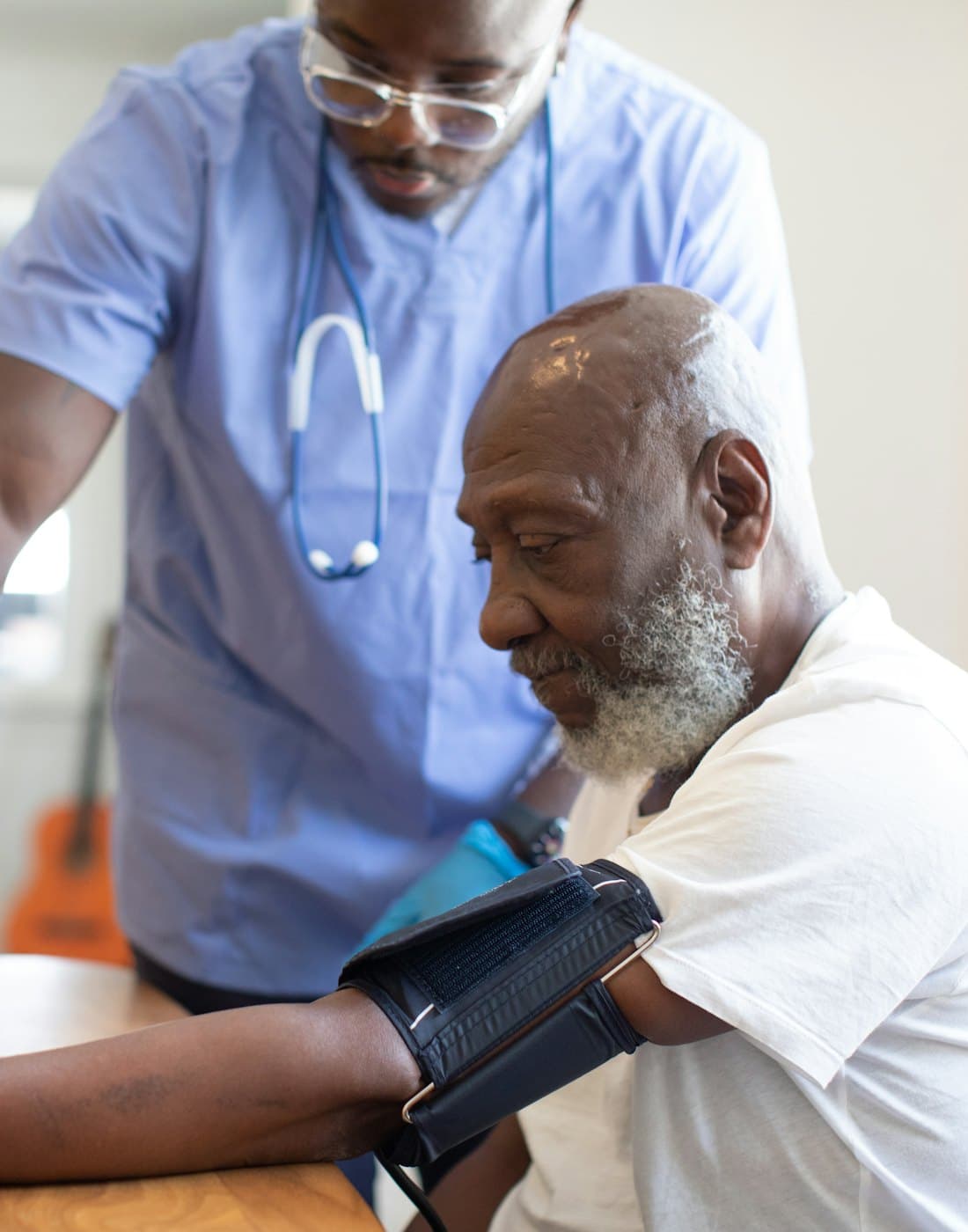 A nurse in scrubs taking an older man's blood pressure at his kitchen table